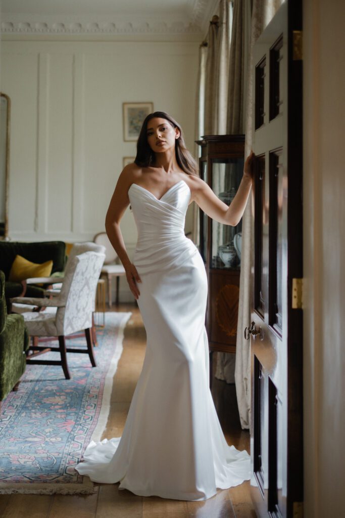 Suzanne Neville Saffron Wedding Dress - A fit-and-flare dress with a tailored bodice and asymmetrical draping at the waist. Featuring a romantic sweetheart neckline and finished with elegant covered buttons for a sophisticated look.