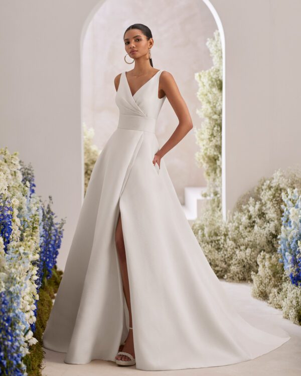 Rosa Clará Taori Wedding Dress - Elegant princess wedding dress crafted in cloqué. Featuring a V-neckline, V-back and straps. The pockets at the skirt, front slit and sash complete the look.